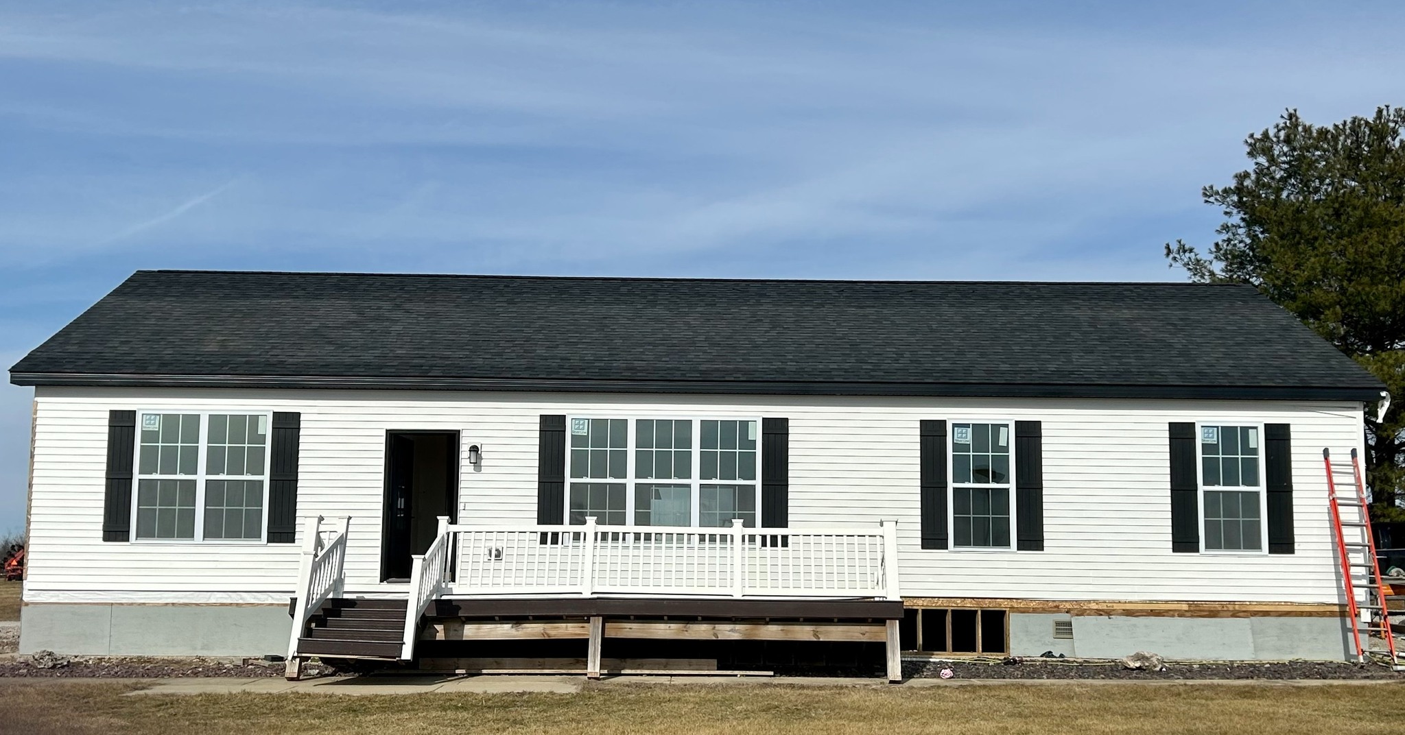 A single-story white house with a dark roof and black shutters. It has a wide front porch with railings and a few steps, set against a clear blue sky.