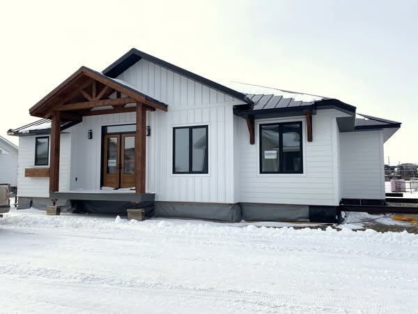 A modern, single-story house with white siding and a gabled roof, featuring a wooden front porch and windows, set against a snowy landscape.