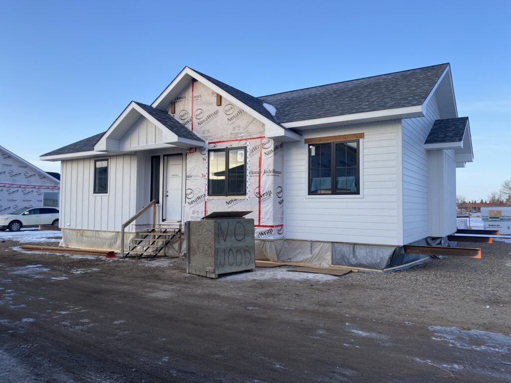 Single-story house under construction with white siding and a partially exposed frame. A box labeled "No Wood" sits in front. The clear sky sets a calm tone.