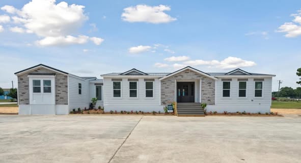 A single-story modular home with a stone facade and white siding sits on a large concrete driveway. Clear blue sky and scattered clouds above.
