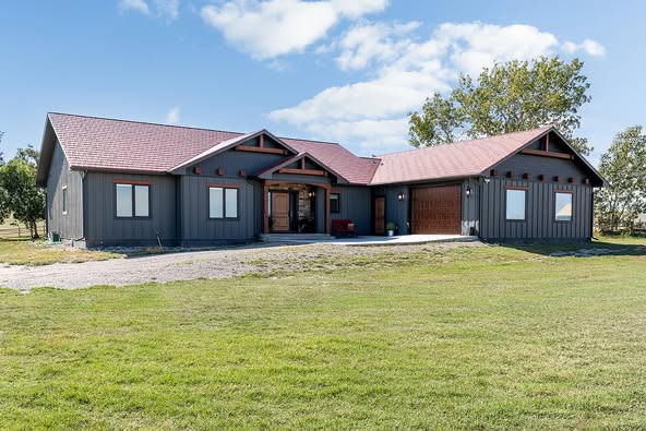 A modern, single-story house with gray siding and red roof sits on a grassy lawn under a blue sky. Large windows and a wooden front door add elegance.