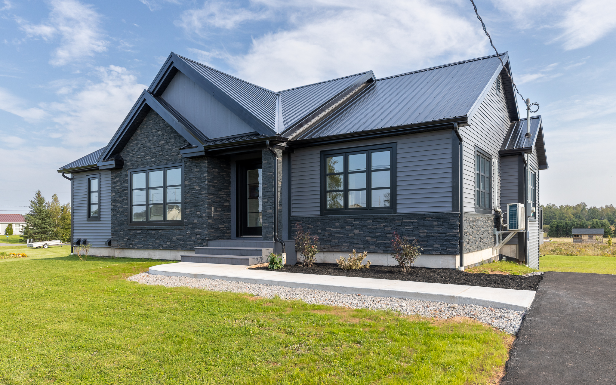 Modern single-story house with dark gray siding and roof, large windows, and a small front porch, set against a bright sky and green lawn.