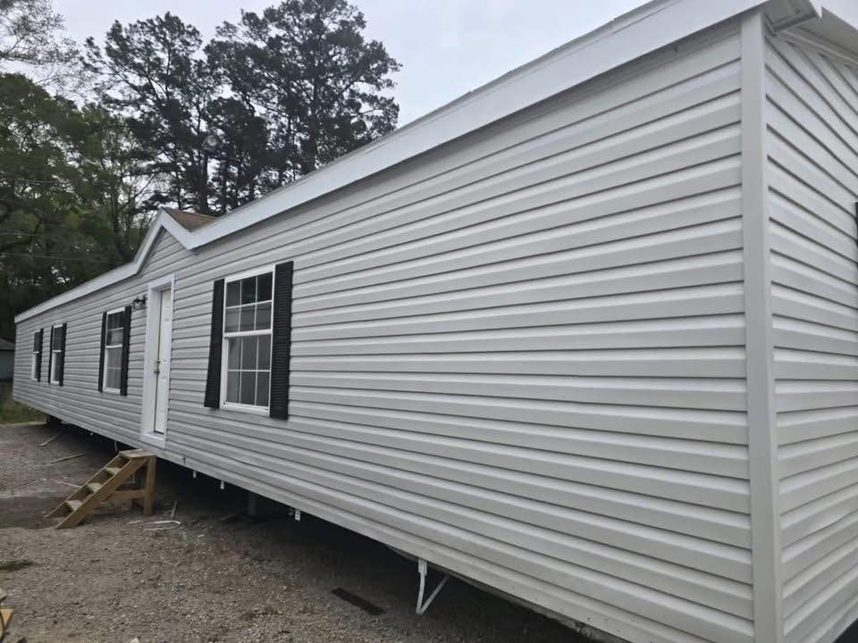 A long, white mobile home with black shutters and small wooden steps at the entrance is set on a gravel area. Tall trees surround the home, creating a serene setting.