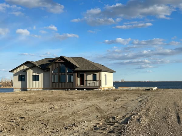 A modern house with large windows stands on a barren plot near a calm body of water, under a blue sky dotted with fluffy clouds, conveying tranquility.