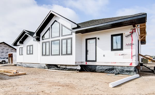 White modern house under construction with large windows and black roof. Unfinished exterior sections expose building materials. Clear sky above.