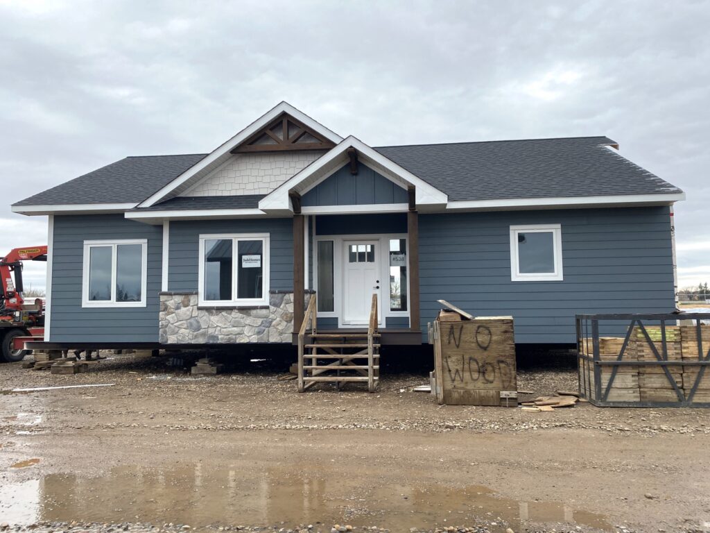 A small blue house under construction on a muddy lot, featuring a gabled roof, stone detailing, and a front porch. A crate labeled "No Woo" is visible.