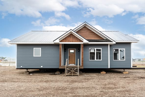 A modern, single-story modular home with gray siding and a metal roof sits on a dirt lot. The facade features a central entrance with a small porch.