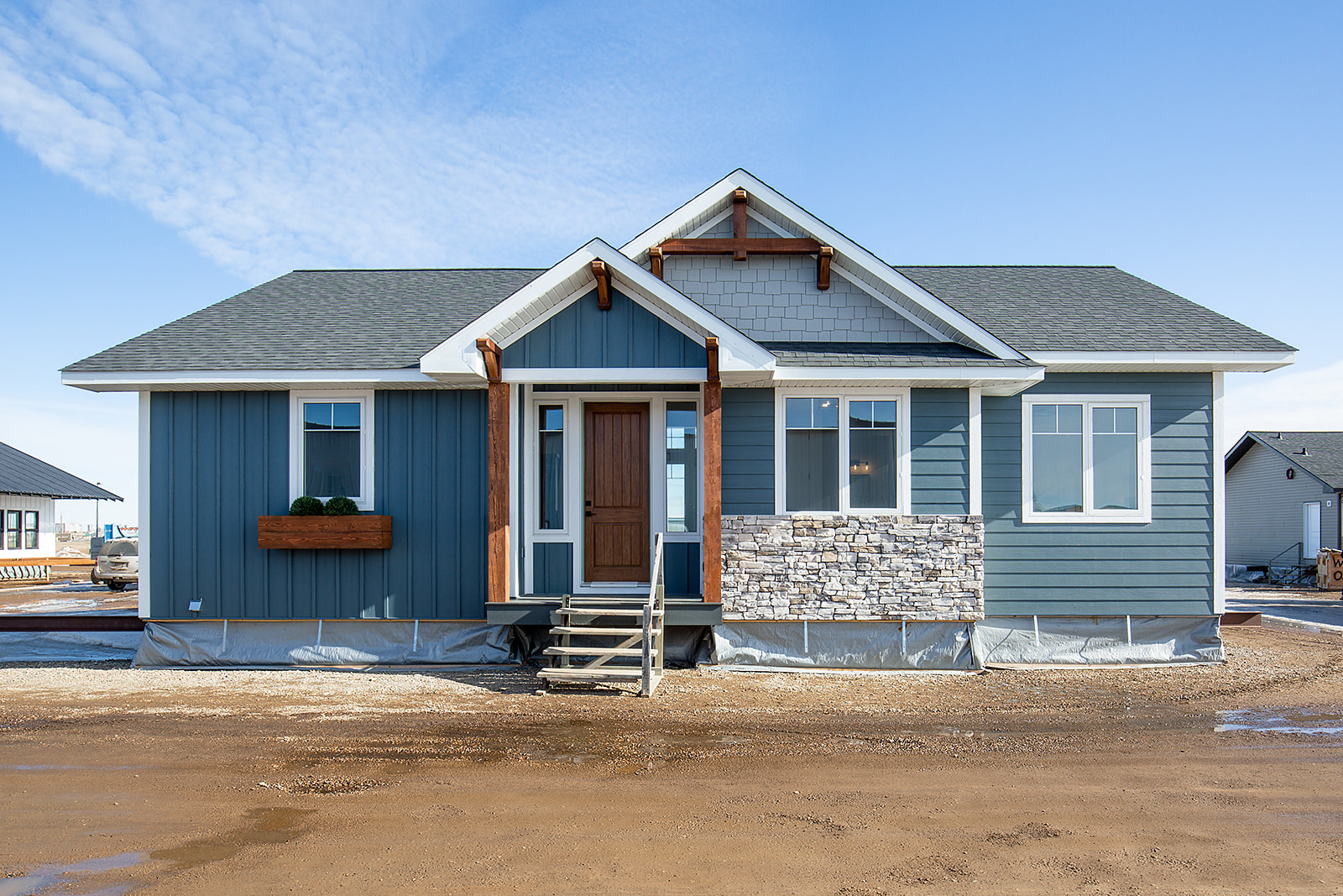 Single-story blue house with a wooden door, gray shingles, and stone accents under a clear sky. White-framed windows and a small front porch add charm.