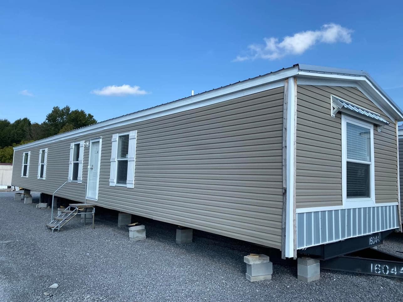 Tan and white mobile home with metal stairs on a gravel lot, under a clear blue sky. Trees are visible in the background, conveying a calm setting.