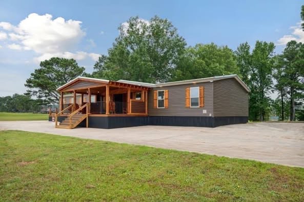 A modern brown modular home with a wooden porch, surrounded by green grass and trees under a blue sky with clouds, creating a serene, inviting scene.