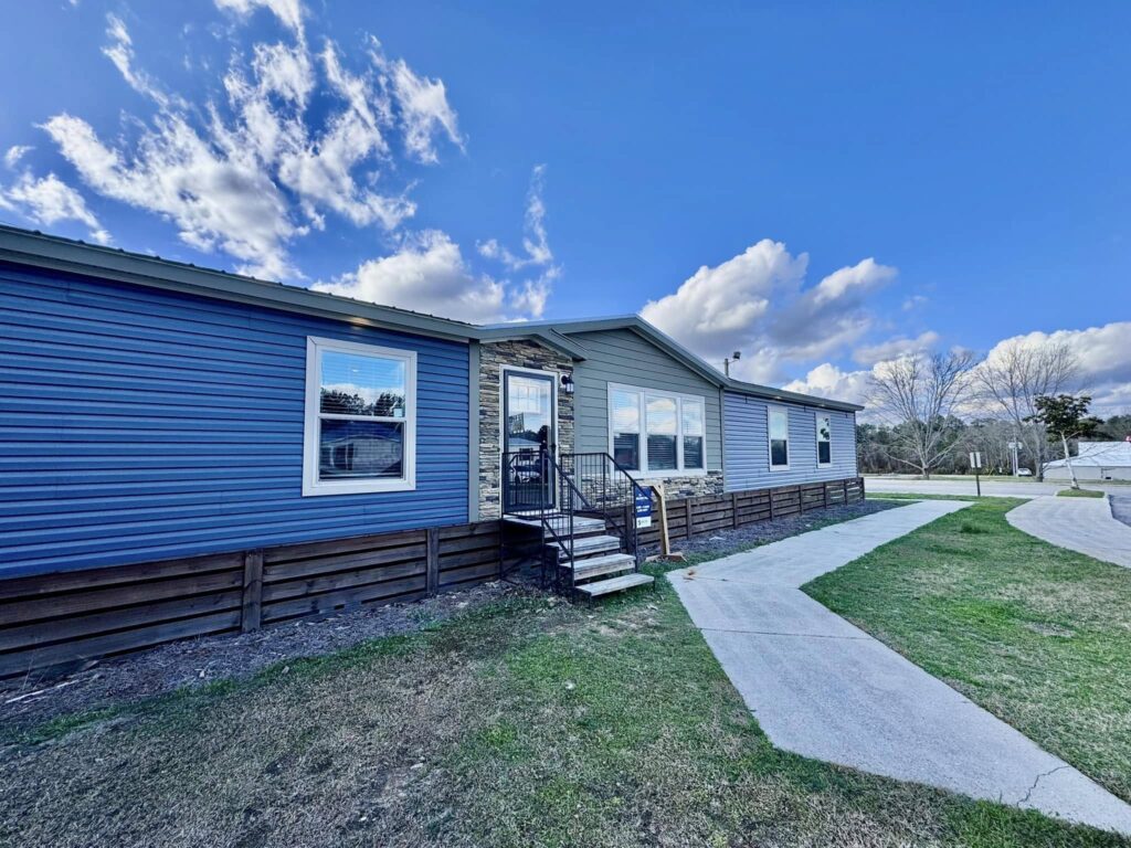 A modern blue and gray manufactured home with a stone accent entryway sits under a vivid blue sky. A curved concrete path leads to the stairs, creating a welcoming feel.