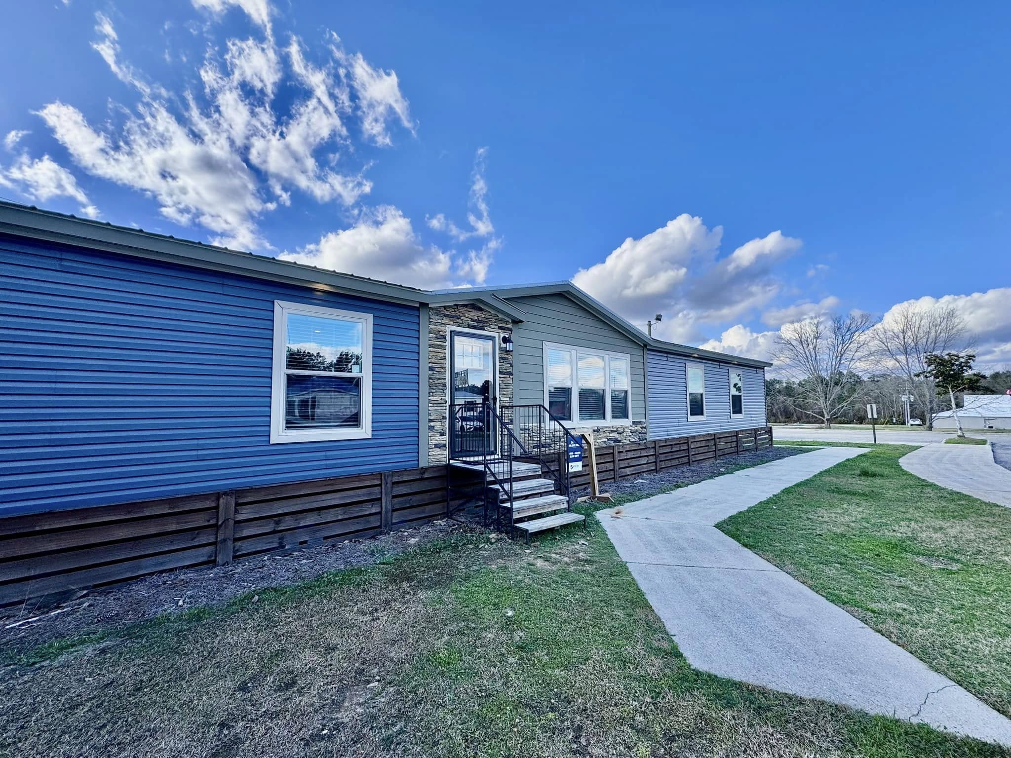 A modern blue and gray manufactured home with a stone accent entryway sits under a vivid blue sky. A curved concrete path leads to the stairs, creating a welcoming feel.