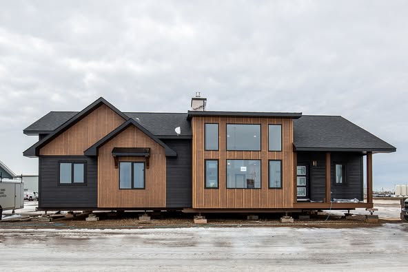 Modern prefabricated home with wood and dark paneling, large windows, and gable roof on a raised platform. Overcast sky, conveying a calm atmosphere.