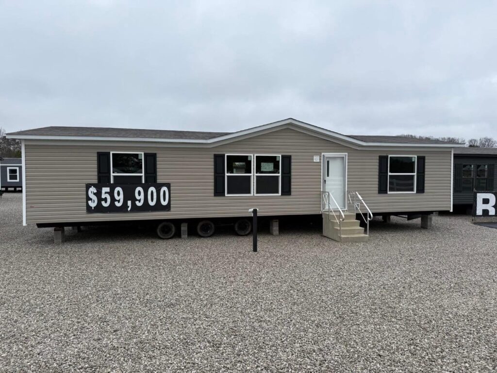 A beige mobile home sits on gravel, priced at $59,900. It has black shutters, large windows, and a small white staircase leading to the door.