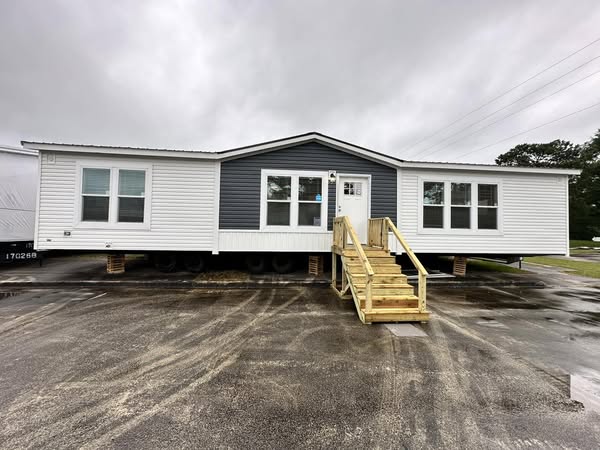 A modern mobile home with white siding and a gray accent wall centered at the entrance. Wooden steps lead to the door. Overcast sky, wet pavement.