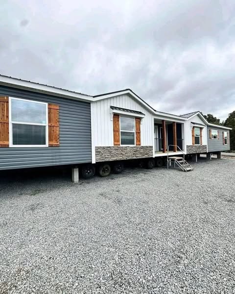 Modern mobile home with blue and white paneling, wood shutters, and stone details on a gravel lot under overcast skies, conveying a rustic ambiance.