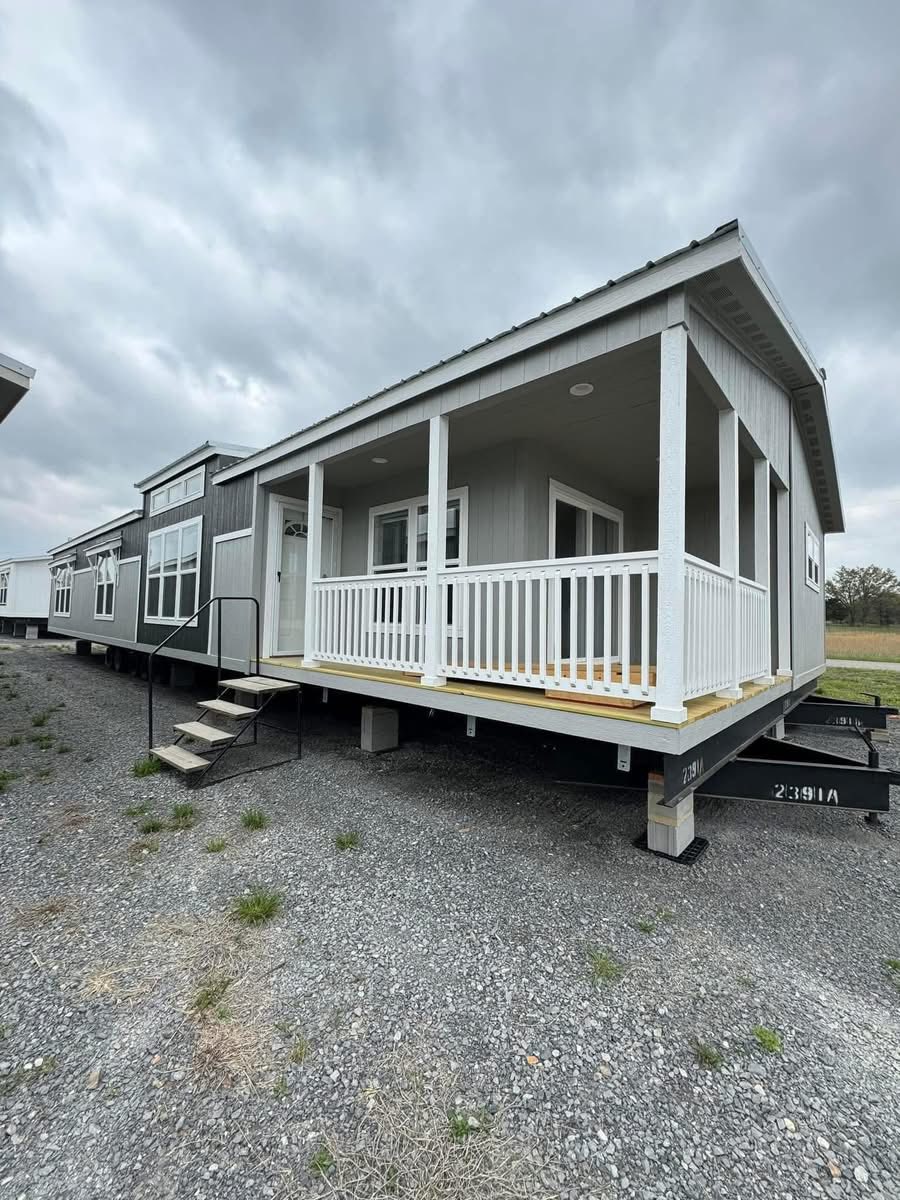 A gray modular home with a front porch and white railing sits on a gravel lot. The cloudy sky and barren surroundings create a subdued atmosphere.