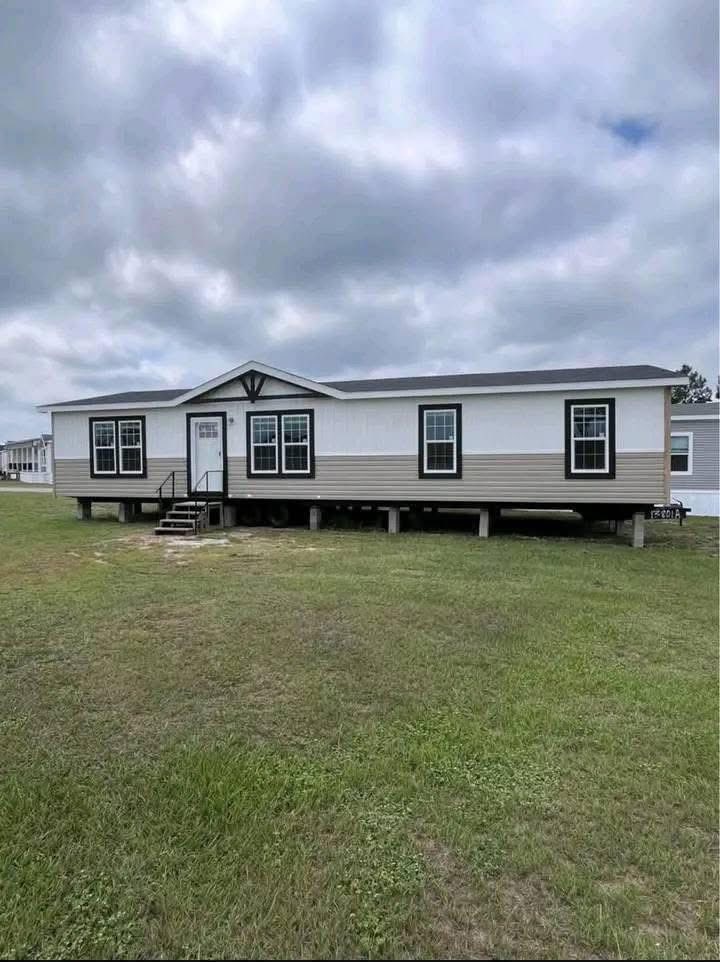 A beige and white manufactured home with multiple windows stands on concrete blocks on a grassy field. Overcast skies create a serene atmosphere.