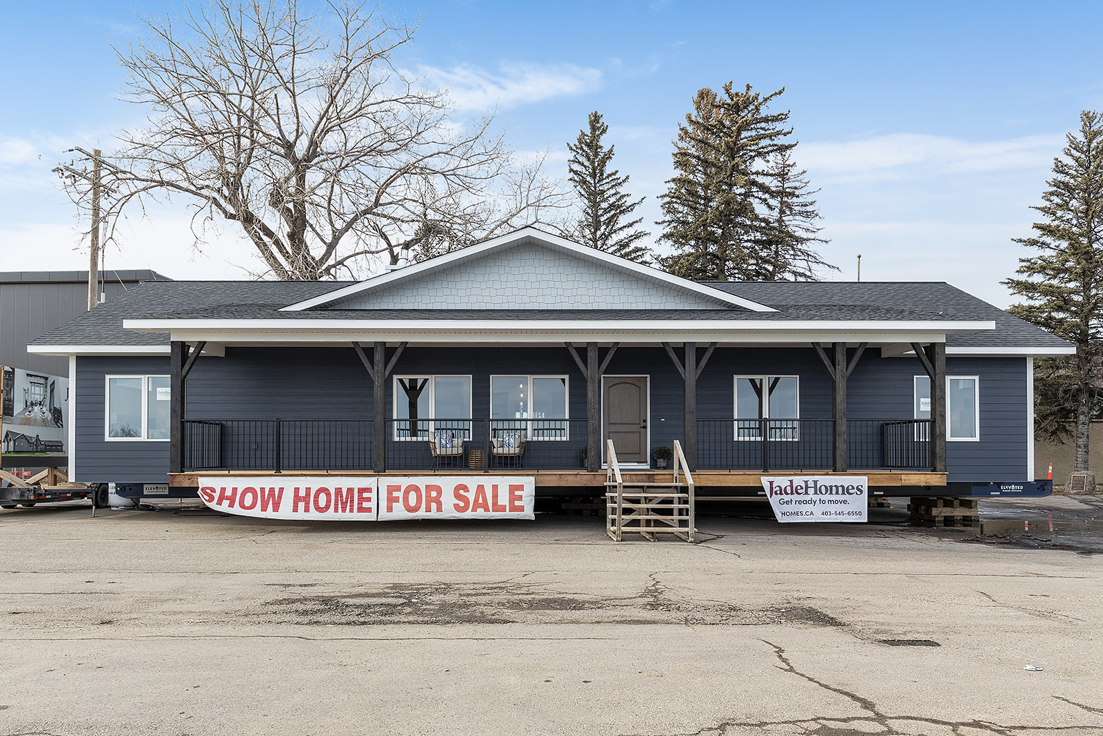 A modern, single-story house with dark blue siding and a gray roof is displayed as a show home for sale. A large banner and signs with contact details are visible. The setting is outdoors with a clear sky and leafless trees in the background.