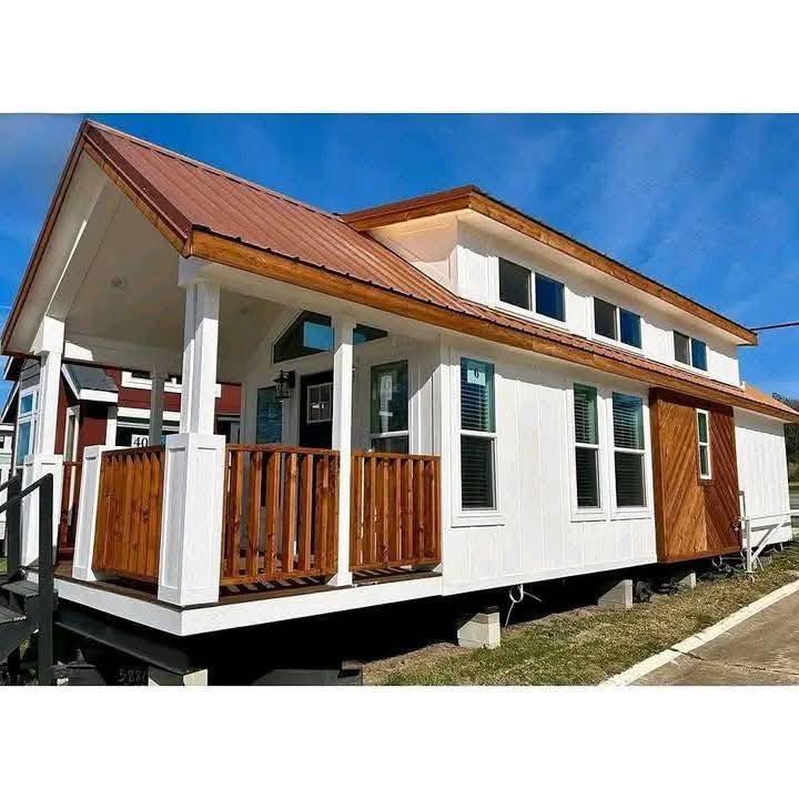 A modern tiny house with a sloped brown roof and a white exterior. It features a wooden front porch, multiple windows, and a welcoming entrance.