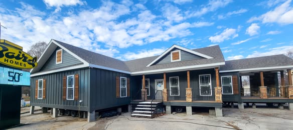 A modern, dark gray modular home with a gabled roof, wooden pillars, and a small front porch under a bright blue sky with wispy clouds.
