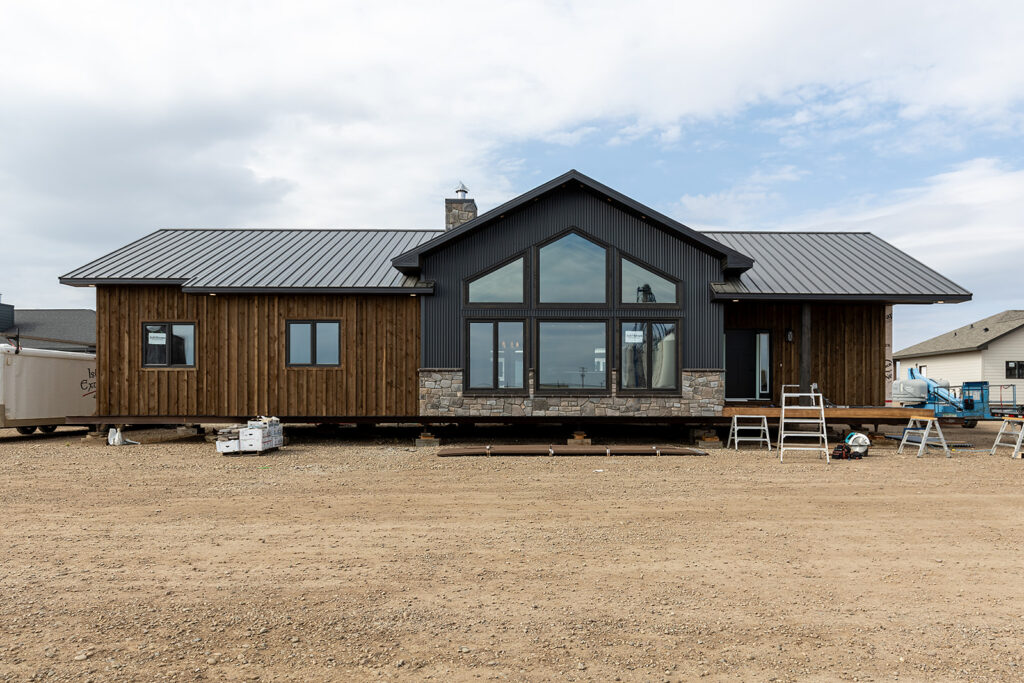 Modern brown and gray house under construction, with large windows and metal roof, set against a cloudy sky. Tools and ladders surround the site.