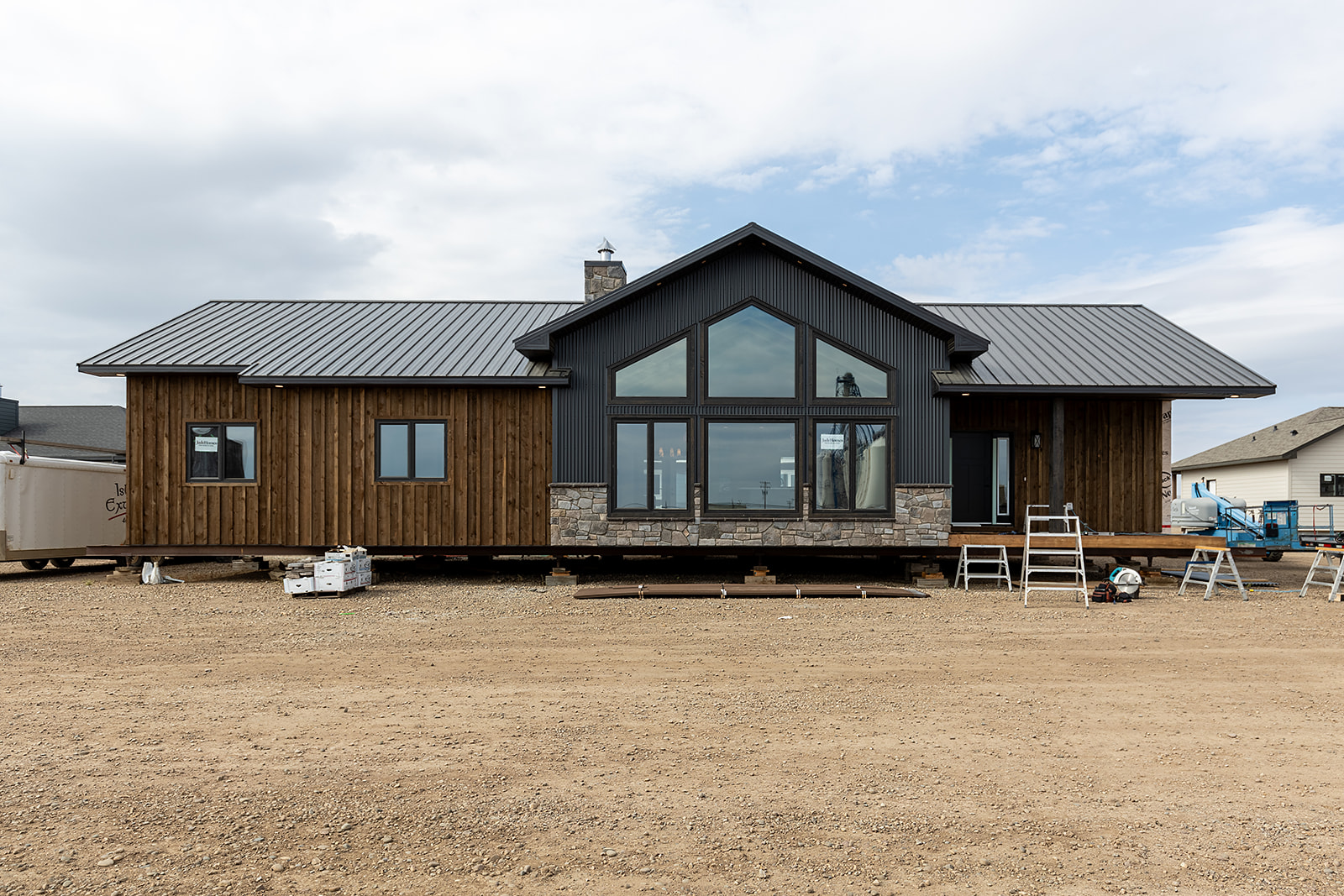 Modern brown and gray house under construction, with large windows and metal roof, set against a cloudy sky. Tools and ladders surround the site.
