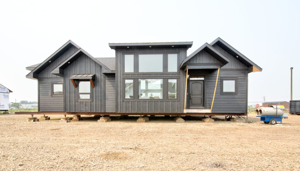 Modern modular home on a construction site, elevated on blocks. Features dark wood siding, large windows, and multiple peaked roofs, under a clear sky.