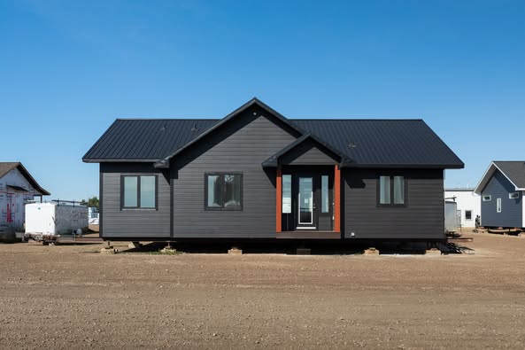 Single-story, dark grey modular house with a black metal roof on an arid dirt lot, clear blue sky, and neighboring homes partially visible.