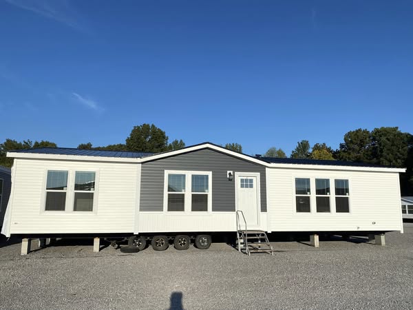 A white and gray mobile home with large windows and a central door is parked on gravel, surrounded by trees under a clear blue sky.