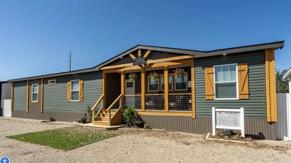 A modern manufactured home with green siding and wooden accents. It features a small porch, two flower baskets, and landscaped plants, set under a clear blue sky.