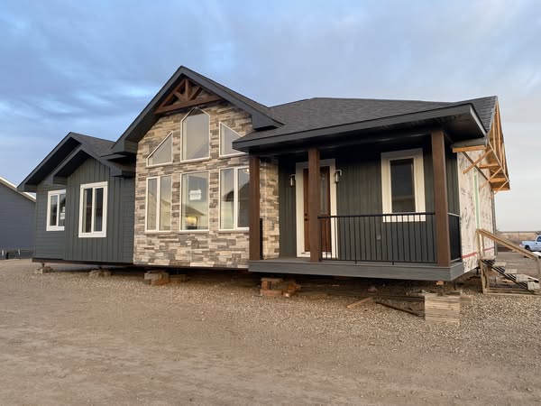 Modern house under construction on a dirt lot, featuring a stone facade, large windows, dark siding, and a small porch with wooden beams.