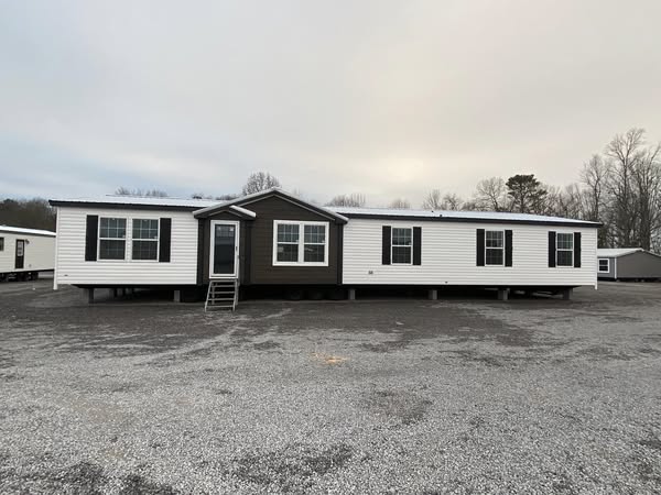 A manufactured home with white siding and dark brown accents, set on a gravel lot. Overcast sky and bare trees create a subdued winter atmosphere.