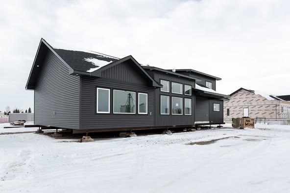 A partially constructed gray house rests on temporary supports in a snowy landscape, surrounded by other structures and building materials under a cloudy sky.