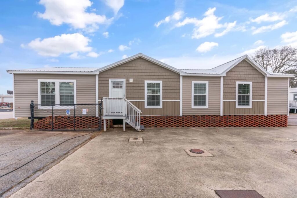 Single-story modular home with tan siding and white trim, featuring a small front porch and multiple windows. Set against a clear blue sky.