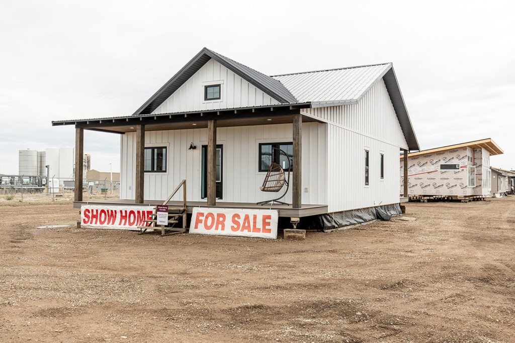 White farmhouse-style show home with a metal roof, on a dirt lot. Large "For Sale" sign, hanging chair on porch. Overcast sky, industrial background.