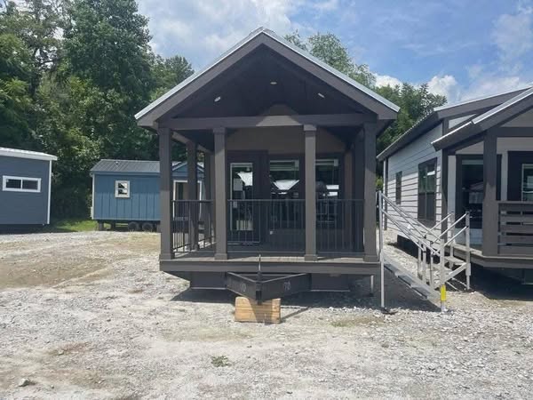 Small, modern tiny house with a covered front porch and dark trim, set on gravel. Surrounded by trees under a blue sky with fluffy clouds, evoking simplicity.