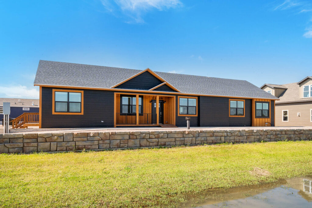 Single-story black house with warm wooden trim and a gabled roof, set on a stone foundation. Bright green lawn under a clear blue sky.