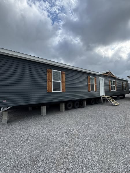 A large gray mobile home on a gravel lot under a cloudy sky. It features wooden shutters, a small staircase at the entrance, and an overcast atmosphere.