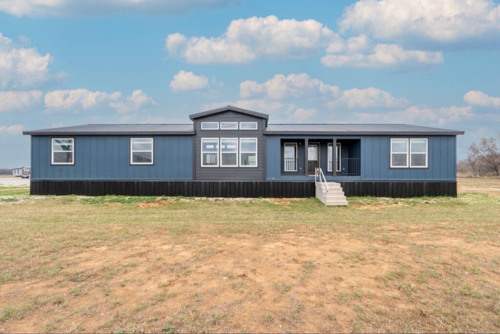 A modern blue mobile home sits on a vast grassy expanse under a cloudy blue sky, featuring large windows and a central porch with white steps.