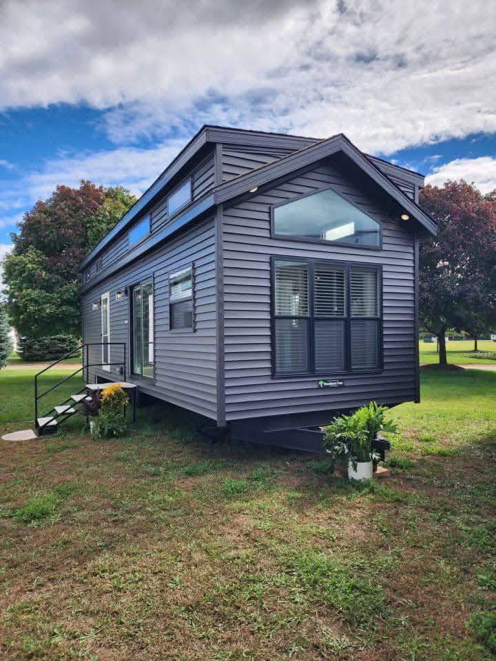 A modern tiny house with dark gray siding and large windows sits in a grassy field. Potted plants adorn the porch steps, creating a cozy, serene vibe.