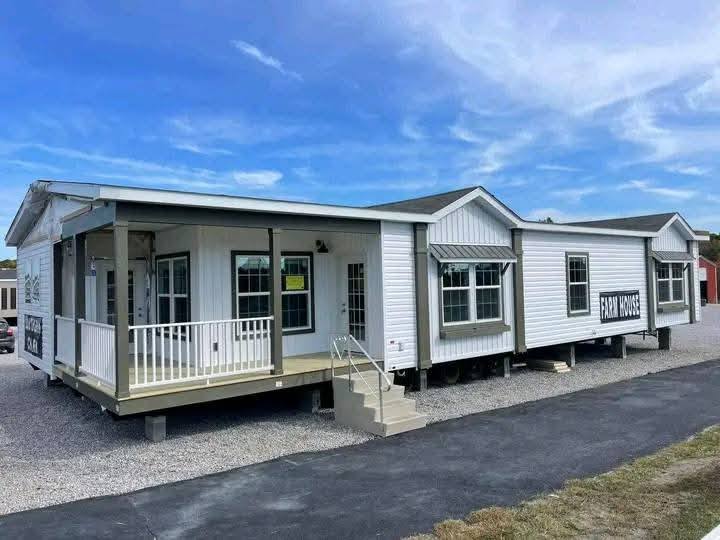 A modern modular home on a gravel lot under a blue sky. Features include a large porch, multiple windows, and "Farm House" signage, giving a cozy feel.