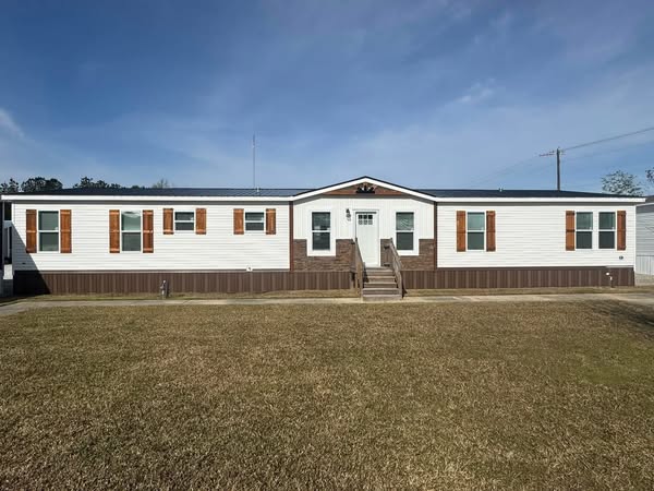 Single-story white manufactured home with brown accents, centered on a lawn under a clear blue sky. The setting is calm and inviting.