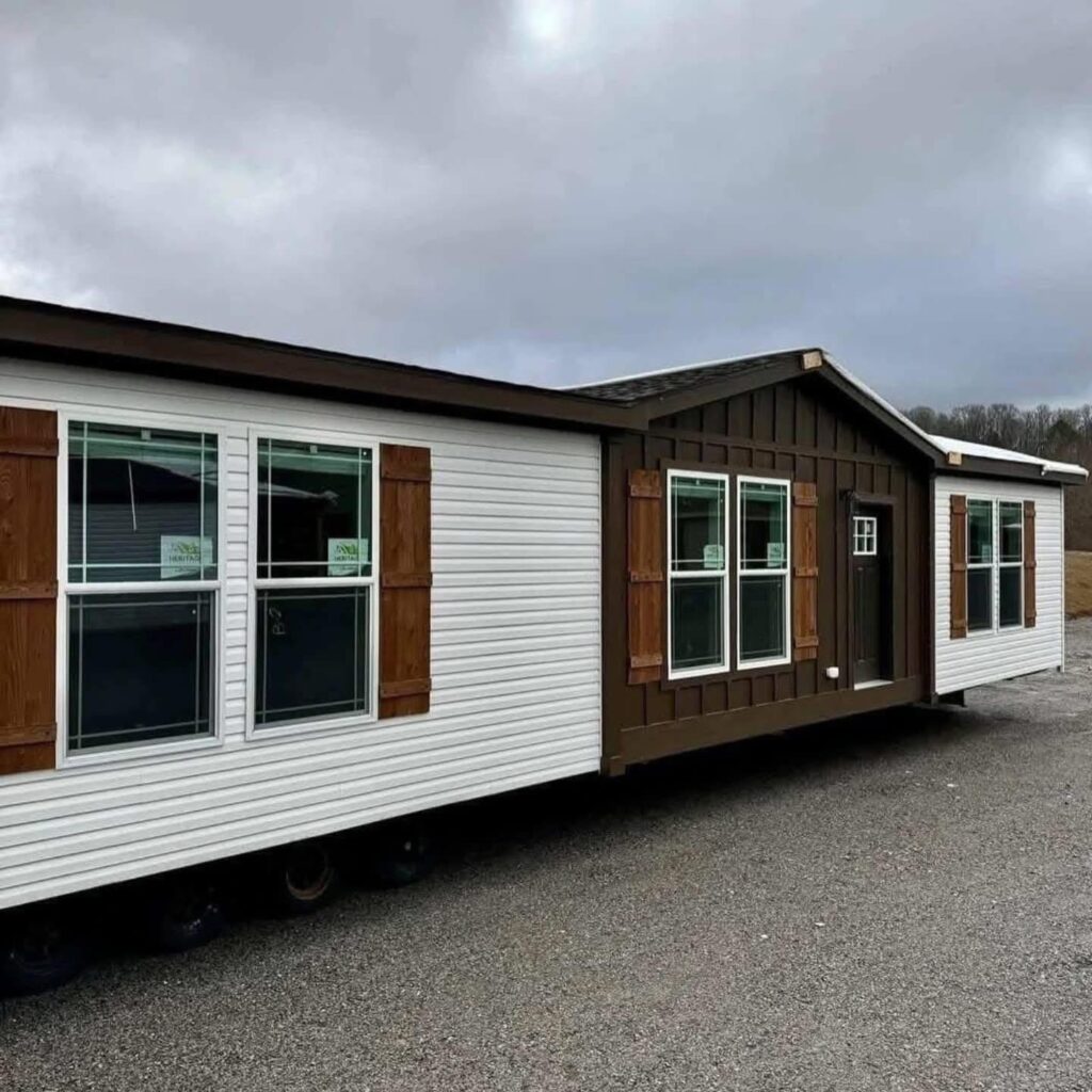 A white modular home with brown accents and wooden shutters sits on a gravel lot under an overcast sky, conveying a simple, rustic feel.
