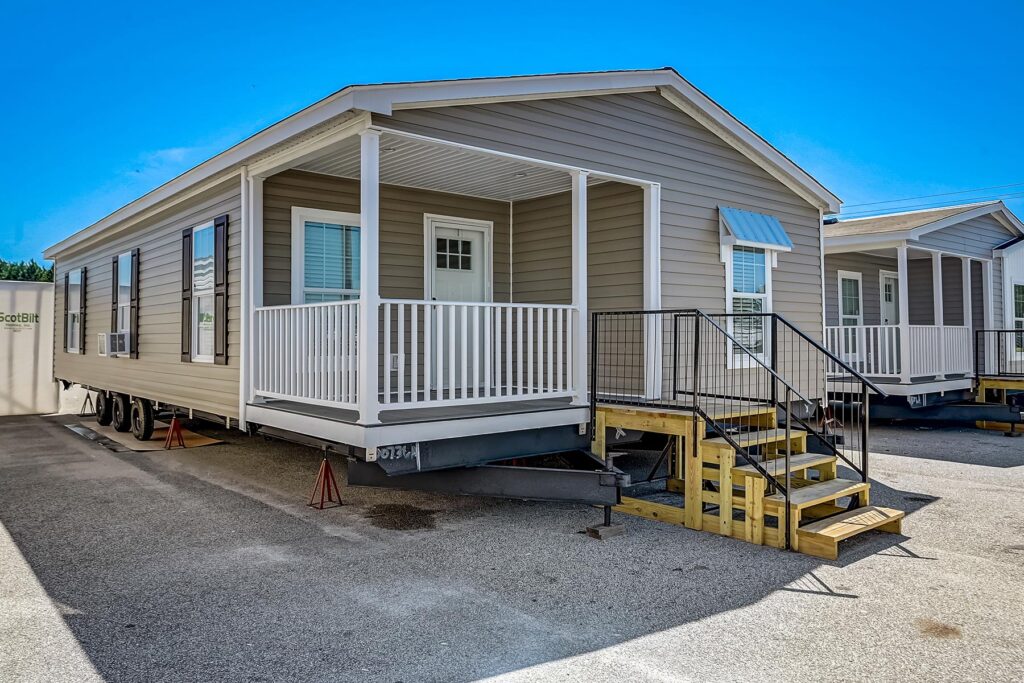 A beige mobile home with white trim, a front porch, and stairs is parked on a lot under a clear blue sky, conveying a clean, new appearance.