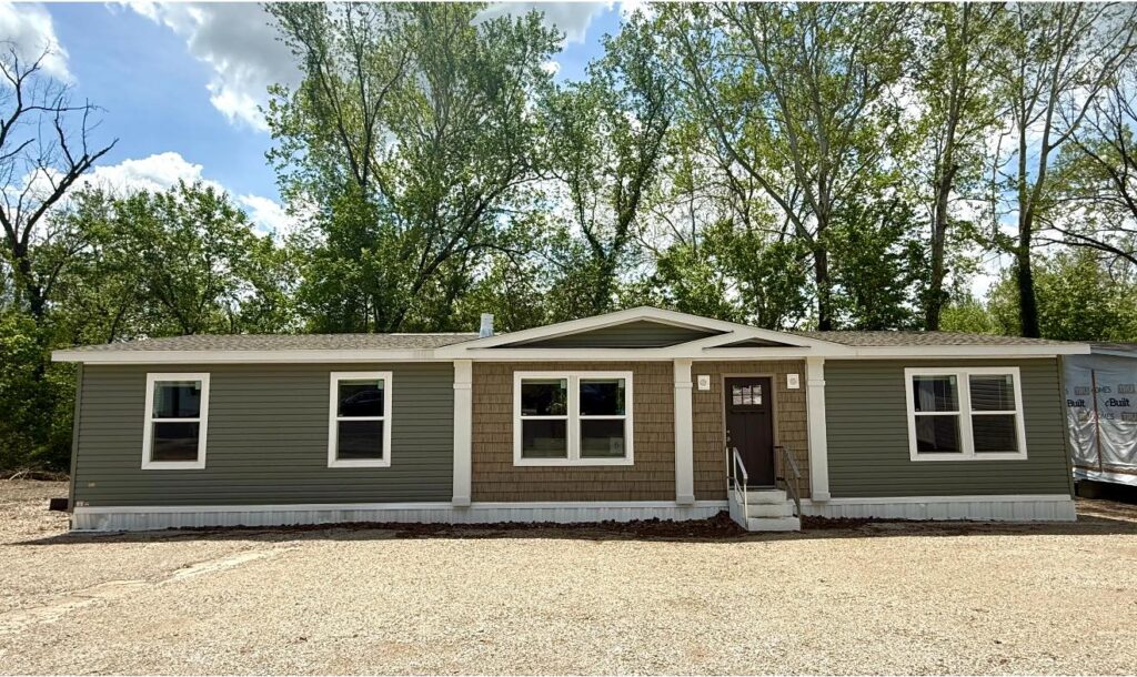 A green, single-story modular home with beige accents sits in front of lush trees under a blue sky. Gravel path leads to a central door. Calm atmosphere.