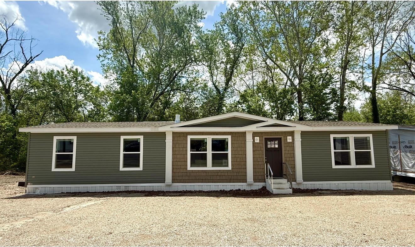 A green, single-story modular home with beige accents sits in front of lush trees under a blue sky. Gravel path leads to a central door. Calm atmosphere.