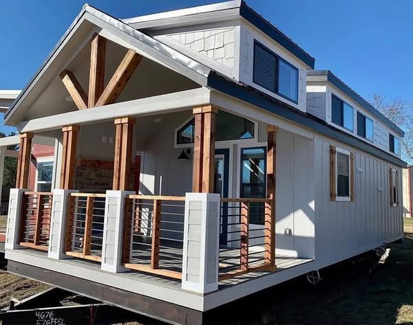 A modern tiny house with a white exterior, wooden trim, and large windows. It features a small porch with railings, set against a clear blue sky.