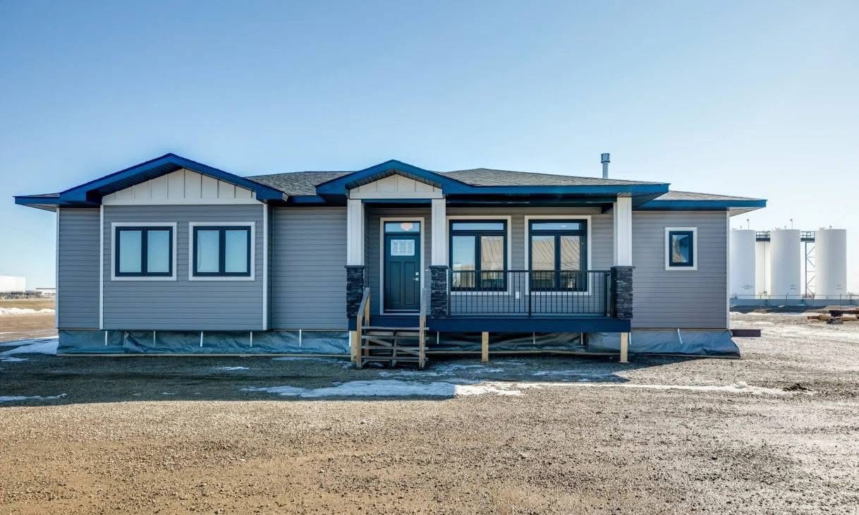 A single-story modular home with light gray siding and a dark gray roof sits in a barren landscape. The front porch with railings is centered. The sky is clear.