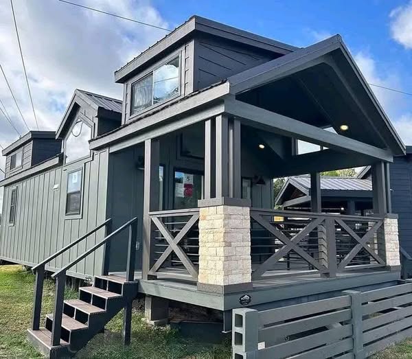 Modern tiny house with a dark gray exterior, featuring a small porch with a wooden railing, stone accents, and a lofted second story under a clear blue sky.